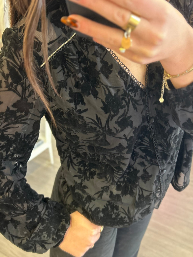Blouse noire en voile transparent avec motifs floraux effet velours et manches longues légèrement volumineuses.